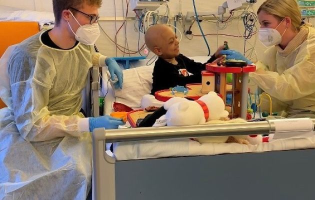 Zwei Erwachsene in Schutzkleidung am Krankenbett eines krebskranken Kindes 